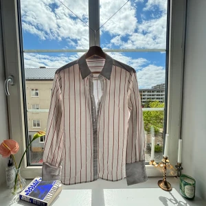 Vit/ randig skjorta från NA-KD - Säljer en vit skjorta från NA-KD med vertikala ränder. Skjortan har klassisk krage, knäppning framtill och långa ärmar. Perfekt för en trendig och stilren look.