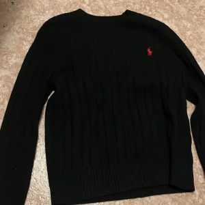 Svart Polo Ralph Lauren - Säljer en svart kabelstickad tröja från Polo Ralph Lauren med klassisk röd logga broderad på bröstet. Tröjan har rund halsringning och långa ärmar, perfekt för en stilren och tidlös look.