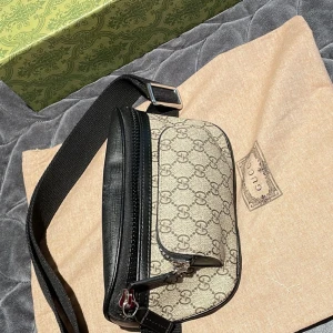 Gucci beige och svart axelväska - Säljer en snygg axelväska från Gucci i beige canvas med klassiskt GG-mönster och svarta skinn-detaljer. Väskan har justerbart band och dragkedja framtill. Perfekt för att bära det viktigaste med stil.