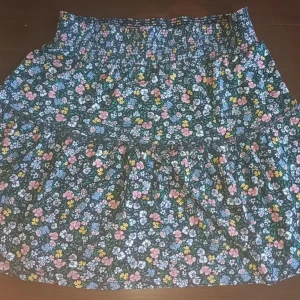 Blommig kortkjol med smock i midjan - Supersöt kortkjol med blommigt mönster i rosa, blått, gult och vitt på mörk botten. Kjolen har smock i midjan och är lätt utställd för en bekväm passform. Perfekt för dig som gillar färg och mönster!