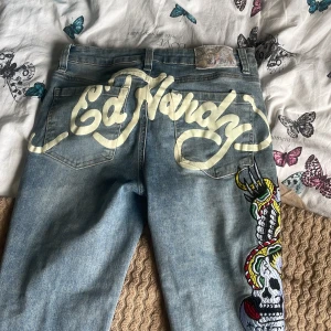 Ed Hardy jeans med tryck och broderi - Säljer ett par ljusblå Ed Hardy jeans med stort tryck bak och färgglatt broderi med döskalle på benet. Byxorna har klassisk femficksdesign och bootcut-modell. Perfekta för dig som gillar statement-plagg och streetstyle.