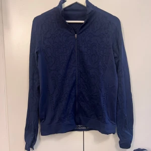 Mörkblå träningsjacka med mönster - Mörkblå träningsjacka med diskret mönster och hel dragkedja framtill. Jackan har lång ärm med tumhål. Står att den är från Crivit, storlek 36/38 S. Helt oanvänd, utan prislapp.