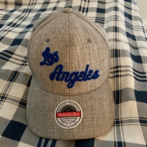 Grå keps från Mitchell & Ness med Los Angeles-brodyr - Säljer en grå keps från Mitchell & Ness med blå broderad text 'Los Angeles' framtill och logga bak. Klassisk böjd skärm och justerbar passform. Perfekt för dig som gillar sportig stil.