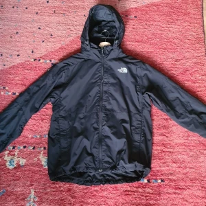Svart vindjacka från The North Face - Säljer en svart vindjacka från The North Face med huva och dragkedja framtill. Jackan har justerbar dragsko i huvan och meshfoder på insidan. Perfekt för blåsiga dagar och enkel att packa ner i väskan.
