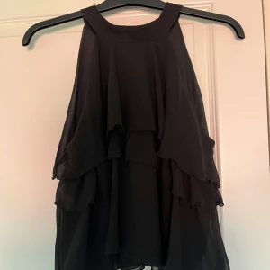 Svart volangig ärmlös blus - Säljer en svart blus med flera lager volanger och hög hals. Blusen är ärmlös och har en elegant, flowy passform som ger en snygg siluett. Perfekt för dig som vill ha något stilrent men ändå med det lilla extra. Blusen är aldrig använd