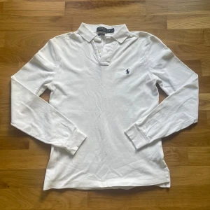 Ralph lauren tröja  - Klassisk vit långärmad pikétröja från Polo Ralph Lauren med blå broderad logga på bröstet. Tröjan har krage, knappar vid halsen och är i ett mjukt bomullsmaterial. Slim fit-modell som passar perfekt till många olika stilar.