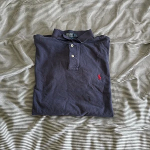 Mörkblå pikétröja från Ralph Lauren - Säljer en klassisk mörkblå pikétröja från Ralph Lauren med den ikoniska röda loggan broderad på bröstet. Tröjan har korta ärmar och en knappslå med två knappar. Perfekt för en stilren och avslappnad look.
