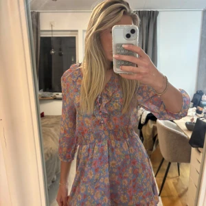 Blommig kortklänning med volangkrage från Zara - Supersöt blommig klänning i blått med rosa och gula blommor från Zara. Klänningen har volangdetaljer vid kragen, knappar framtill och trekvartsärm. Smockad midja ger en fin passform och kjolen är lätt utställd.