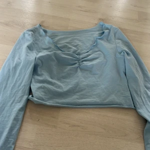 Ljusblå långärmad topp - Säljer en ljusblå långärmad topp med rynkad detalj vid bysten. Toppen har en v-ringad design och är perfekt för en stilren look. Passar bra till både jeans och kjol.