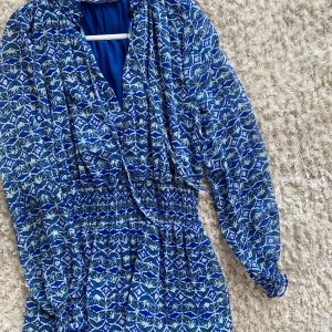 Blå mönstrad klänning från Zara - Säljer en blå mönstrad klänning från Zara med v-ringning och långa ärmar. Klänningen har ett elastiskt midjeband som ger en smickrande passform. Perfekt för en färgglad look! Knappt använd och så fin!💙