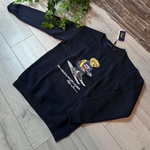 Svart polo bear hoodie  - Säljer en svart tröja från Ralph Lauren med ett coolt tryck av en björn som åker skidor. Tröjan är långärmad och tillverkad i 100% bomull, vilket gör den både bekväm och stilren. Perfekt för dig som vill ha något unikt i garderoben!