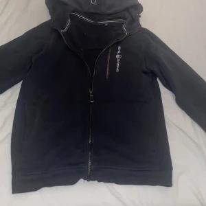 Svart zip hoodie med dragkedja från Sail Racing - Snygg svart zip från Sail Racing med dragkedja och huva. Perfekt för sommar kvällar när de blir lite kylligare 