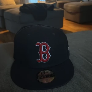 Svart keps med röd broderad bokstav - Snygg svart keps från New Era med en stor röd broderad bokstav på framsidan. Kepsen är en officiell on-field cap och har en MLB-logga på baksidan. Perfekt för sportiga tillfällen eller som en stilren accessoar.