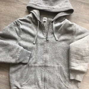 Grå hoodie från GAP - Säljer en mysig grå hoodie från GAP med dragkedja och fodrad med mjuk fleece. Perfekt  våriga dagar med sin varma huva och praktiska fickor. Passar både för avslappnade dagar och utomhusaktiviteter.
