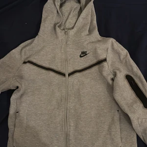 Grå hoodie nike tech fleece  - Säljer en grå hoodie från Nike med dragkedja och huva. Jackan har svarta detaljer och en ficka med dragkedja på ärmen. Perfekt för en avslappnad stil.