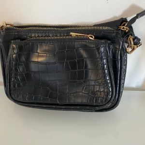 Svart axelväska med krokodilmönster - Snygg svart axelväska med krokodilmönster och guldfärgade dragkedjor. Väskan har flera fack och en justerbar axelrem med en liten rund väska fäst vid remmen. Perfekt för att hålla ordning på dina saker med stil.