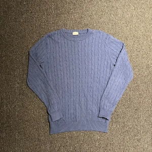 Blå kabelstickad tröja från John Henric - Säljer en snygg blå kabelstickad tröja från John Henric. Tröjan är tillverkad i 100% bomull och har en klassisk rund halsringning. Perfekt för kyligare dagar med sin stilrena design och bekväma passform.