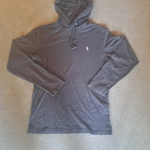 Grå hoodie från Ralph Lauren - Säljer en stilren blå hoodie från Ralph Lauren med en broderad logga på bröstet. Tröjan har långa ärmar och en klassisk huva med dragsko. Perfekt för en avslappnad look. Den är tunn.