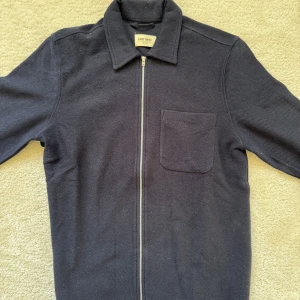 Mörkblå cardigan jacka från East West - Säljer en stilren mörkblå cardigan jacka från East West. Jackan har en dragkedja framtill och en praktisk bröstficka. Perfekt för en avslappnad look. Storlek S