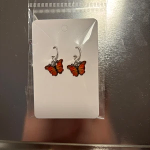 Örhängen med fjärilar - Charmiga örhängen med färgglada fjärilar i orange och svart. Perfekta för att lägga till en lekfull touch till din stil. De hänger från en silverfärgad krok och är lätta att bära. (Billigt material) 
