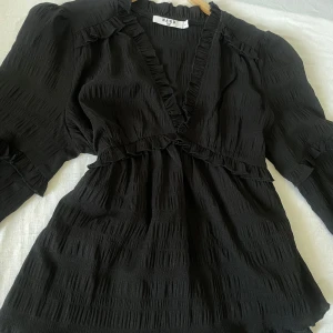 Svart blus från NA-KD - Snygg svart blus från NA-KD med volangdetaljer och v-ringning. Blusen har långa ärmar och en lätt textur i tyget som ger en elegant look. Perfekt för att klä upp eller ner beroende på tillfälle.