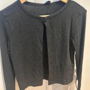 Mörkgrå kofta - Säljer en stilren mörkgrå kofta med rund halsringning och långa ärmar. Perfekt för lager-på-lager-stil och passar till många olika outfits. Prislappen sitter kvar
