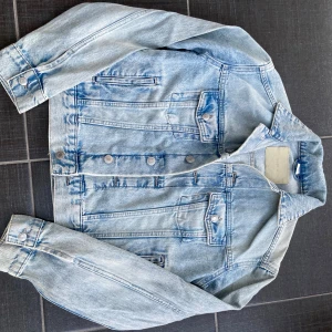 Ljusblå jeansjacka - Säljer en klassisk ljusblå jeansjacka med knappar framtill och två bröstfickor med lock. Jackan har en normal passform och långa ärmar. Perfekt för vår och sommar! Säljer pga att den är för liten. Jackan är i storlek xs-s💗