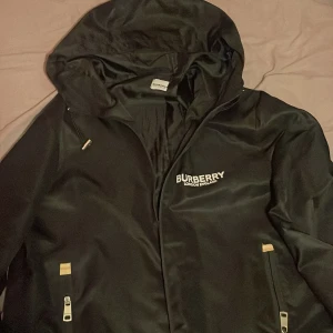 Svart vindjacka från Burberry - Snygg svart vindjacka från Burberry med dragkedja och huva. Jackan har Burberry-logga på bröstet och dragkedjor på fickorna. Perfekt för en stilren look.