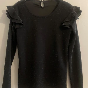 Svart glittrig tröja med volangdetaljer - Snygg svart tröja med glittrigt material och volangdetaljer vid axlarna. Tröjan har långa ärmar och en rund halsringning med en liten öppning bak. Perfekt för en kväll ute eller en festlig tillställning.