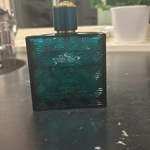 Versace Eau de Toilette - Elegant flaska i mörkgrön glas med Versaces ikoniska Medusa-motiv. Flaskan har en gyllene kork och rymmer 60/70 ml av doften. Perfekt för den som vill ha en lyxig och stilren parfymupplevelse.