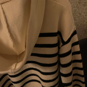 Randig hoodie i beige och svart - Säljer en snygg randig hoodie i beige och svart. Den har en klassisk design med huva och ribbade muddar vid ärmslut och nederkant. Perfekt för en avslappnad stil.