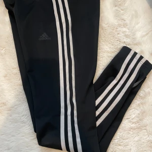 Svarta leggings från Adidas - Snygga svarta leggings från Adidas med de klassiska tre ränderna längs sidorna. Perfekta för träning eller en avslappnad dag. De har en hög midja och är tillverkade i ett stretchigt material för bästa komfort.