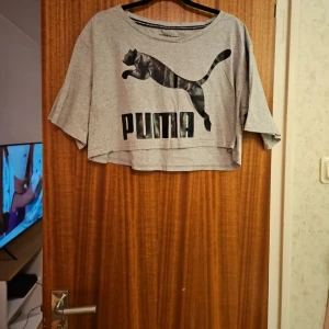 Grå croppad topp från Puma - Säljer en grå croppad topp från Puma med deras ikoniska logga framtill. Toppen har en lös passform och trekvartsärmar, perfekt för en avslappnad stil. Tillverkad i mjukt bomullsmaterial.