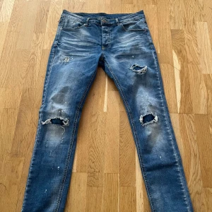 Blå slitna jeans från Kings Will Dream - Säljer ett par blå jeans från Kings Will Dream med slitna detaljer och hål på knäna. Jeansen har en normal passform och är tillverkade i denim. Perfekta för en avslappnad stil.