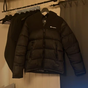 Svart dunjacka från Champion - Säljer en svart dunjacka från Champion med dragkedja och huva. Jackan har en stilren design med Champion-loggan på bröstet. Perfekt för kyligare dagar. Lite fläckar på bröstet under loggan