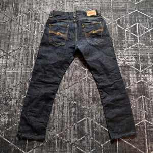 Mörkblå jeans från Nudie Jeans - Snygga mörkblå jeans från Nudie Jeans med klassisk femficksdesign och kontrasterande orange sömmar. Jeansen har en knappgylf och en stilren passform som passar perfekt till vardags. Märkets signaturbroderi syns på bakfickorna.