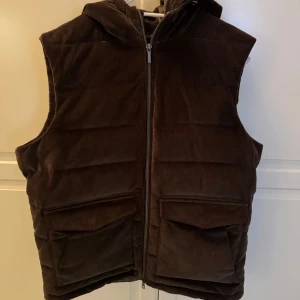 Manchester dunväst från Stenströms - Snygg  mörk grön Manchester dunväst från Stenströms med dragkedja och huva. Perfekt för kyliga dagar när du vill hålla stilen. Västen har praktiska fickor framtill och en quiltad design som ger extra värme. Storlek M. Färgen är alltså mörkgrön/olivgrön, inte brun som den kan tyckas vara på bilderna. 