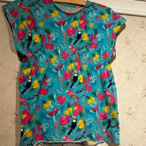 Färgglad blommig topp från Mywear - Säljer en färgglad topp från Mywear med tropiskt mönster av blommor och fåglar. Toppen har en lös passform och är ärmlös, perfekt för varma dagar. Den livfulla designen gör den till ett härligt tillskott i garderoben. STORLEK=140cl