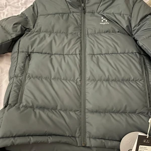 Grön dunjacka från Haglöfs - Säljer en stilren och varm grön dunjacka från Haglöfs. Jackan har en dragkedja framtill och är perfekt för kyliga dagar. Den är designad för att hålla dig varm och bekväm med sin pufferstil.