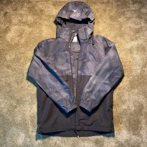 Grön McKinley Skaljacka - Säljer en grön vindjacka från McKinley med huva och dragkedja. Jackan har ett diskret rutigt mönster och elastiska ärmslut. Perfekt för blåsiga dagar! På bilden ser den grå ut men i verkligheten är den lite mer åt det grönare hållet. Jackan är även Namnad men går lätt att stryka över. 10000MM vattenpelare, vind och vattentät, justerbar i nederkant, avtagbar huva. Dragkedja i sidofickoe