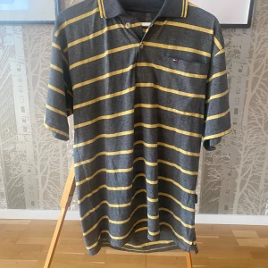 Grå och gul randig pikétröja från Tommy Hilfiger - Säljer en stilren grå pikétröja med gula ränder från Tommy Hilfiger. Tröjan har korta ärmar och en klassisk krage med knappar. Perfekt för en avslappnad look.