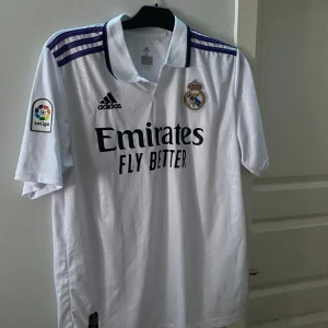 Vit Benzema Real Madrid fotbollströja - Säljer en vit Real Madrid fotbollströja från Adidas med korta ärmar. Tröjan har klubbens emblem och sponsorlogga på framsidan samt spelarnamnet 'Benzema' och nummer 9 på baksidan. Står XL som storlek men passade bra på mig som har M.