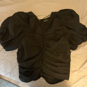 Svart blus med puffärmar - Snygg svart blus från Gina Tricotmed puffiga ärmar och rynkade detaljer framtill. Perfekt för en stilren look. Blusen har en v-ringad design och är tillverkad i ett lätt material.