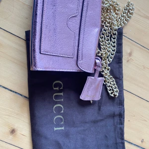 Rosa metallic axelväska från Gucci - Säljer en snygg rosa metallic axelväska från Gucci med guldfärgad kedja och låsdetalj. Väskan har en praktisk ficka på baksidan och en liten nyckelväska. Perfekt för att ge din outfit en lyxig touch.