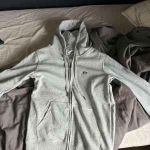 Grå hoodie från Lacoste - Säljer en stilren grå hoodie från Lacoste med dragkedja och den klassiska krokodilloggan på bröstet. Tröjan har långa ärmar och en praktisk huva med snörning. Perfekt för en avslappnad look. Den var medium men har krympt i tvätten