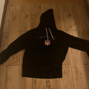 Svart hoodie från GANT - Säljer en svart hoodie från GANT med en stor logga och ett sköldmärke på bröstet. Hoodien har en klassisk design med dragsko i huvan. Perfekt för en avslappnad stil.