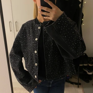 Svart jeansjacka med diamanter  - Snygg svart jeansjacka med diamanter  och knappar framtill. Jackan har en normal passform och långa ärmar, perfekt för en stilren look.