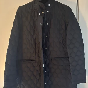 Svart quiltad jacka från GANT - Säljer en stilren svart quiltad jacka från GANT. Jackan har en dragkedja och knappar framtill samt två fickor. Perfekt för kyligare dagar.