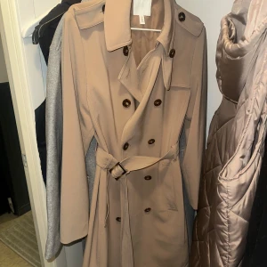 Beige trenchcoat med bälte - Snygg beige trenchcoat med klassisk design och knappar framtill. Jackan har ett bälte i midjan för en figursydd passform och långa ärmar. Perfekt för övergångsperioder.