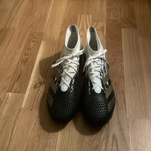 Svartvita adidas predator fotbollsskor - Ett par Adidas predator 20.3. Snygga fotbollsskor från Adidas i svart och vitt med guldiga detaljer. Skorna har snörning och en unik design med dobbar för optimalt grepp på planen. Perfekta för fotbollsspelare som vill ha både stil och funktion.